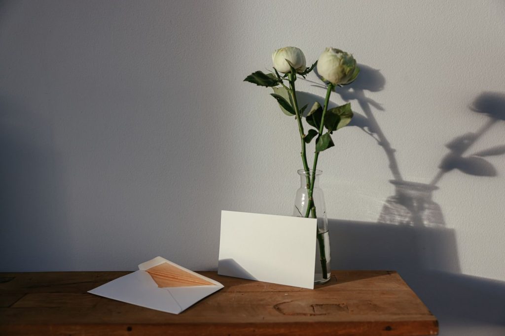 Weiße Rosen in einer Glasvase neben einer leeren Karte und Umschlägen auf einem Holztisch